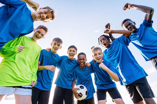 Group of teenage soccer players in blue jerseys on a green field hugging after scorring a goal