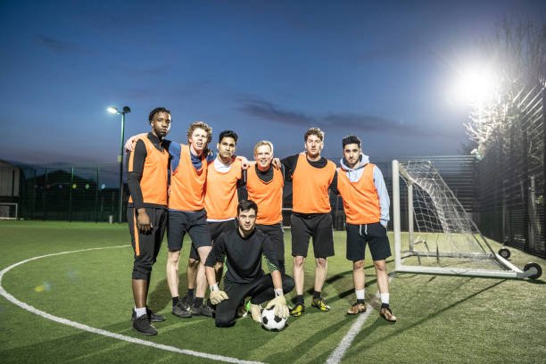 Footballers in orange bibs looking at camera on a floodlit pitch at night, goalkeeper kneeling on floor at front with football